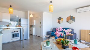 A modern kitchen with white cabinets, stainless steel appliances, and pendant lighting. There is a dining area with a wooden table set for two, and a blue sofa with decorative orchids. - Le Grand Large by Interhome (Sainte Maxime)