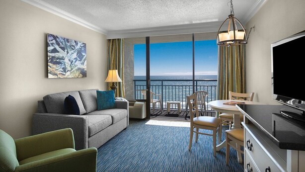 A hotel room with a balcony, a sofa, a dining table, and a TV.