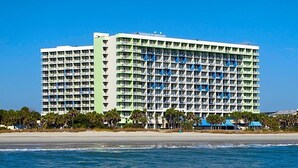 A large hotel with multiple floors and balconies, located near a beach with palm trees.