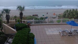 A beach view from a balcony with tiled flooring, a few lounge chairs, and a palm tree.