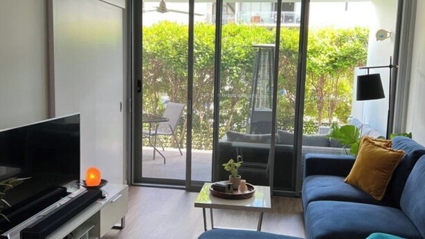 A modern living room with a flat-screen TV, a sofa, a coffee table, and a sliding glass door leading to an outdoor area with a patio chair and a table.