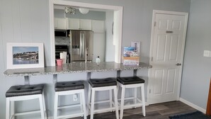 A kitchen with a granite countertop, white cabinets, and a stainless steel refrigerator. There are three bar stools and a framed picture on the counter.