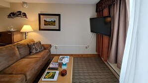 Un salon avec un canapé marron, une table basse avec des livres et une télécommande, un tableau encadré au mur et un téléviseur fixé au mur.