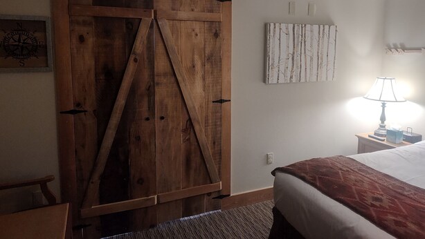 Une chambre avec une porte de grange en bois, un lit recouvert d’une couverture rouge, une table de chevet avec une lampe et un tableau accroché au mur.