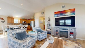 A spacious living room with a large TV, comfortable seating, a kitchen in the background, and a view of the outdoors. - Private Heated Pool, Pet Friendly, Fire Pit (Port Aransas)