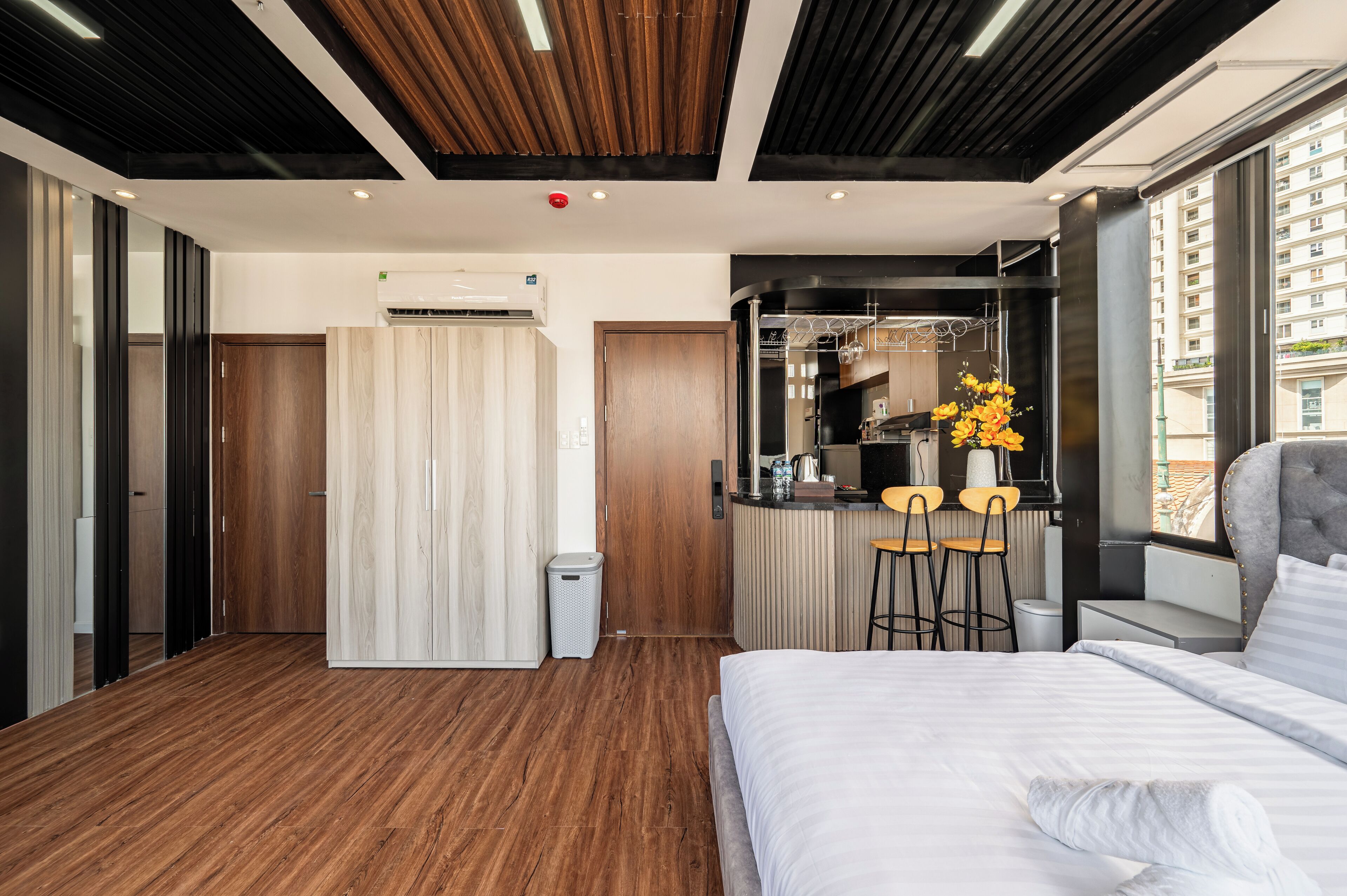 A modern hotel room with a wooden floor, a large bed, a small kitchenette, and a view of the city.