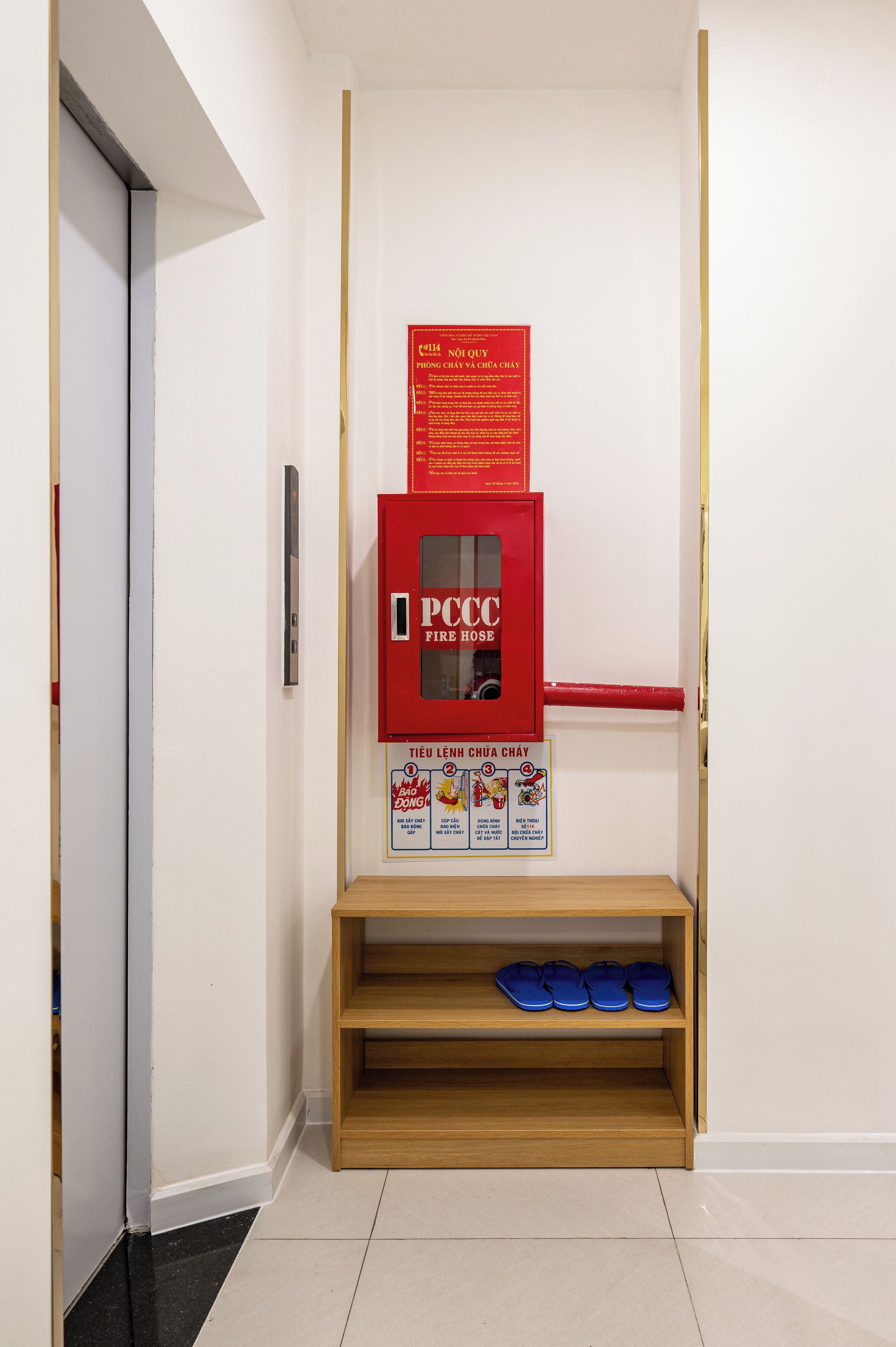 Un couloir avec une armoire rouge contenant un tuyau d’incendie et une étagère en bois où sont rangées des chaussures bleues.