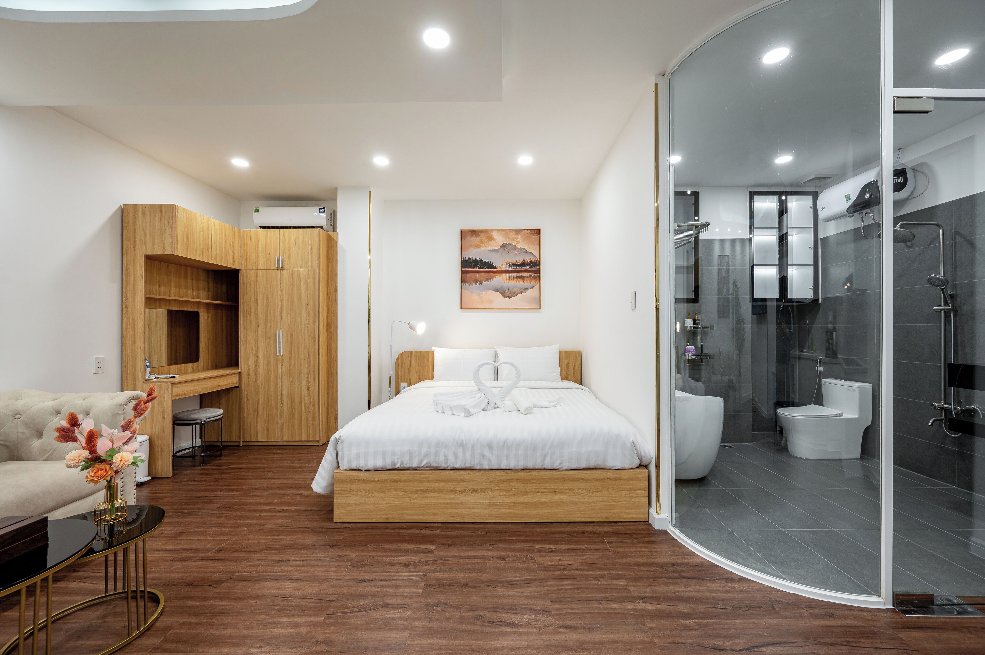 A modern bedroom with a wooden bed, a white sofa, a glass-enclosed shower, and a painting on the wall.