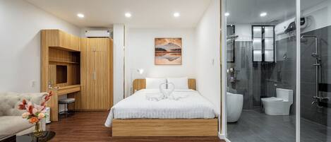 A modern bedroom with a wooden bed, a white sofa, a glass-enclosed shower, and a painting on the wall.