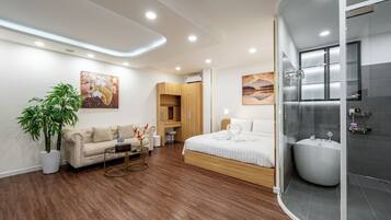 A modern hotel room with a wooden floor, a sofa, a bed, a large potted plant, and a glass-enclosed shower area.
