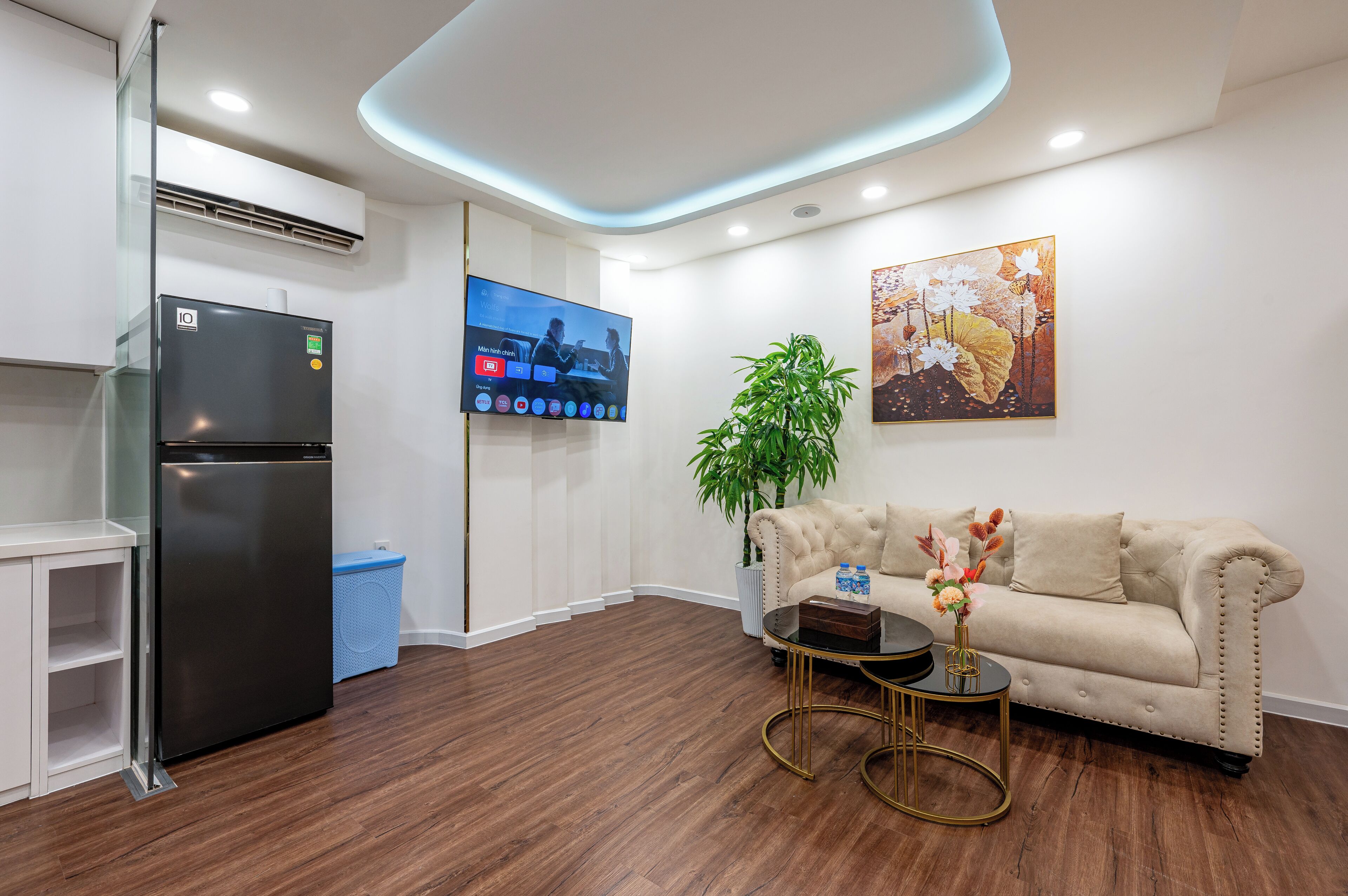 A modern living room with a black refrigerator, a beige sofa, a small round table with a vase, and a large flat-screen TV mounted on the wall.