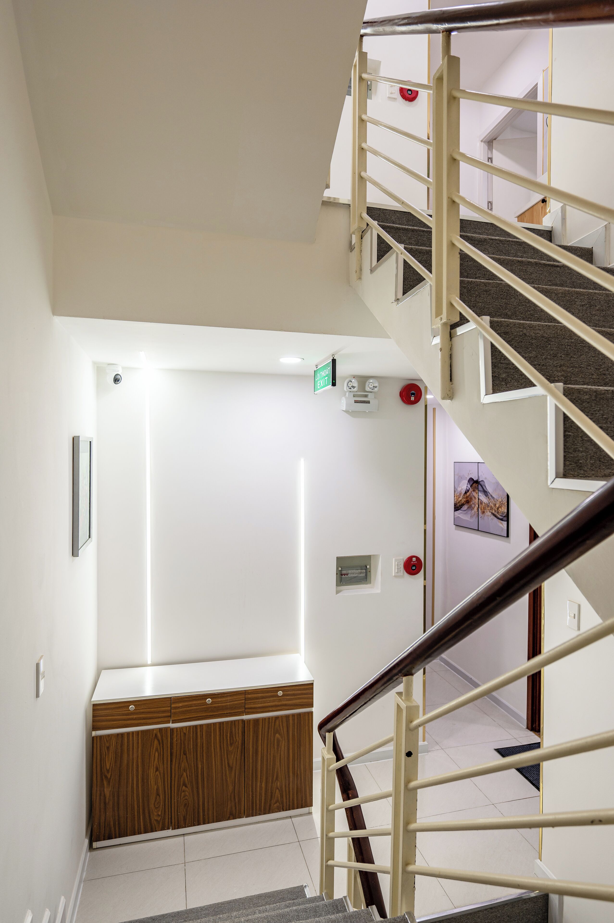 Uma escada com corrimão, parede branca, um banco de madeira e uma porta com placa de saída de emergência.