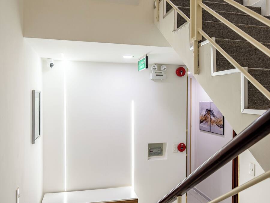 Uma escada com corrimão, parede branca, um banco de madeira e uma porta com placa de saída de emergência.