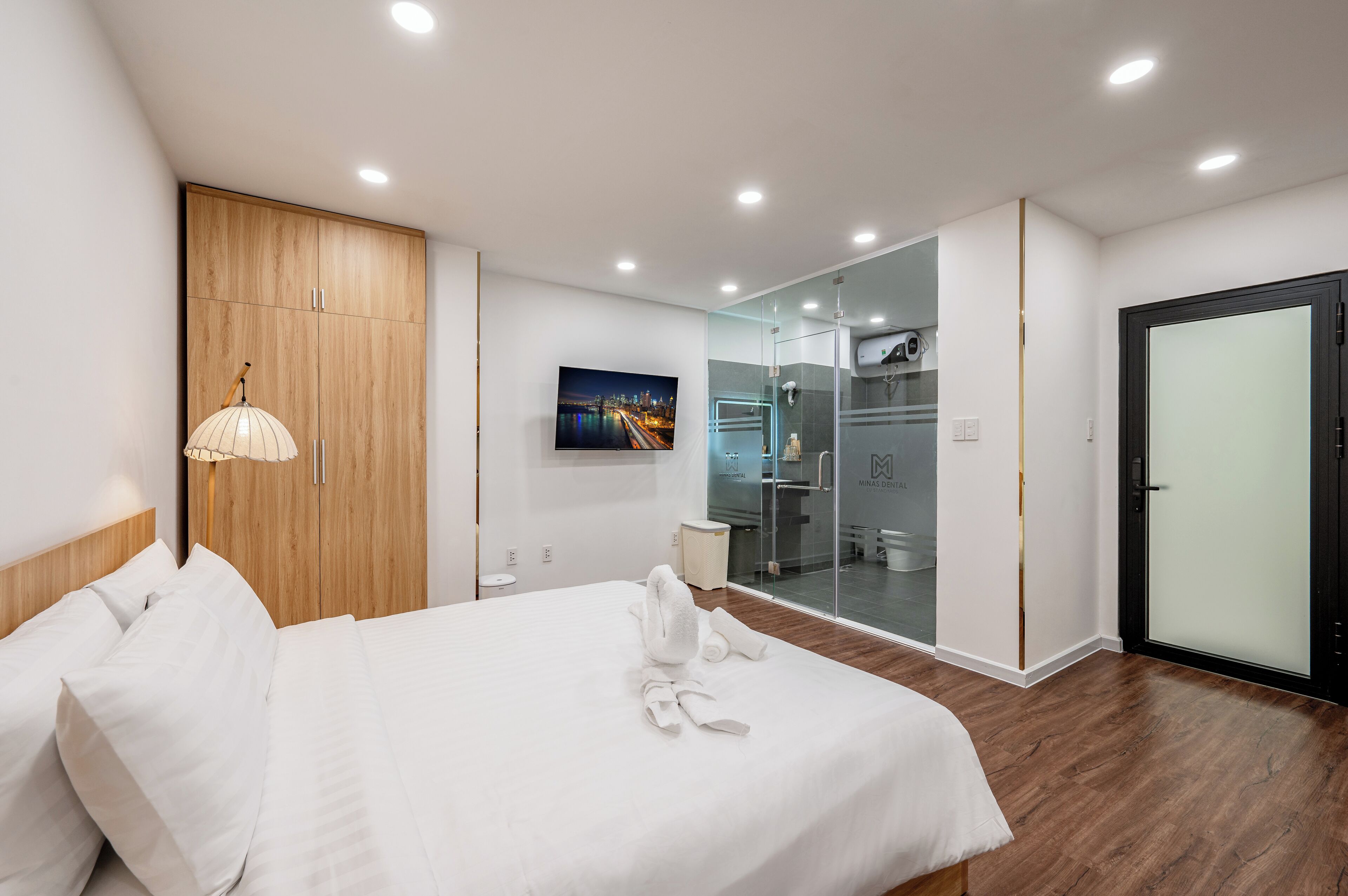 A modern hotel room with a large bed, a wooden wardrobe, a glass-enclosed shower area, and a cityscape view on the wall.