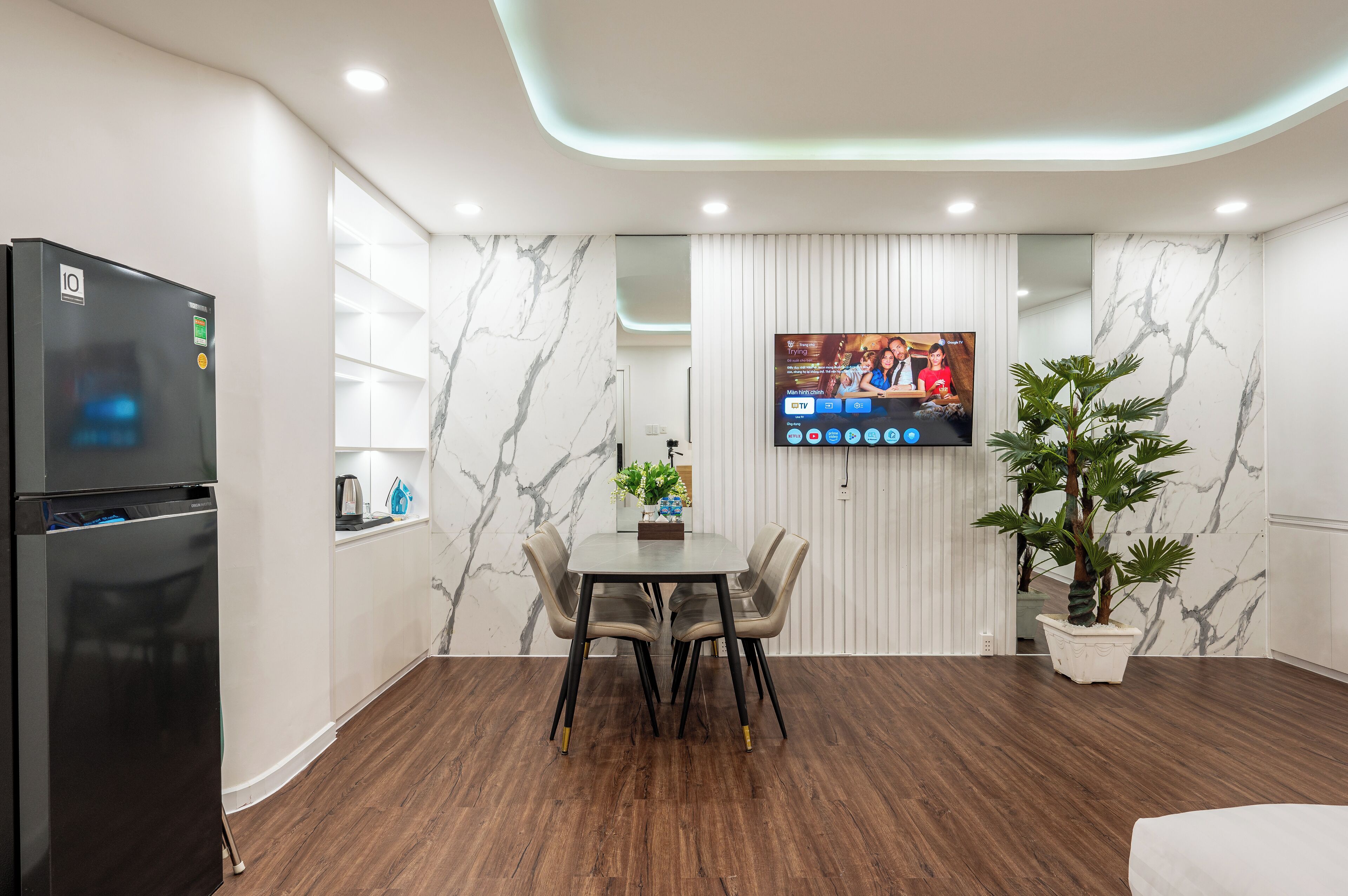 A modern kitchen with a built-in refrigerator, a dining table with chairs, a TV mounted on the wall, and a potted plant.