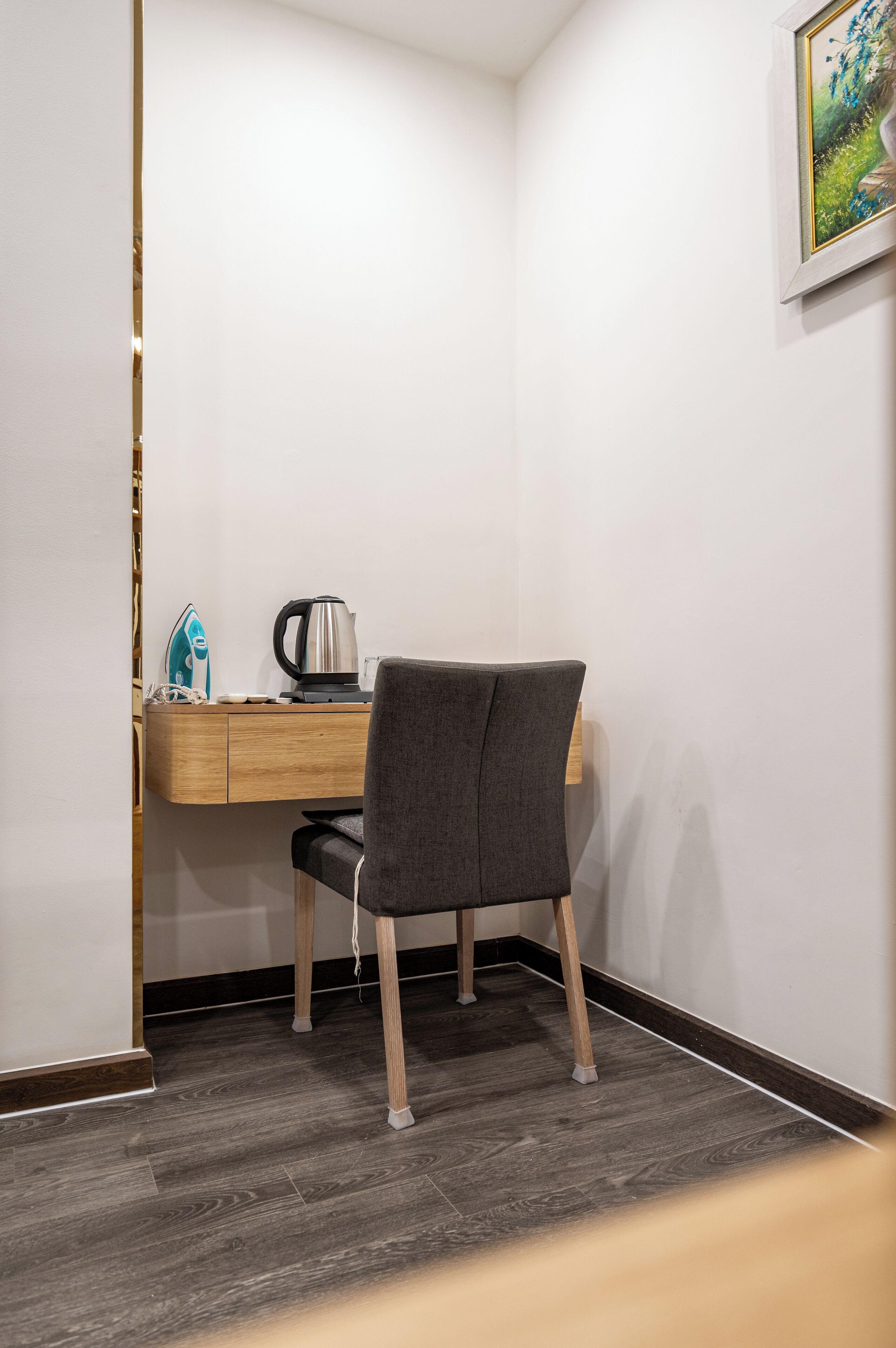 A corner room with a wooden desk, a chair, a kettle, and a framed picture on the wall.