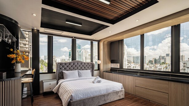 A modern bedroom with a large bed, a desk, and a view of the cityscape through large windows.