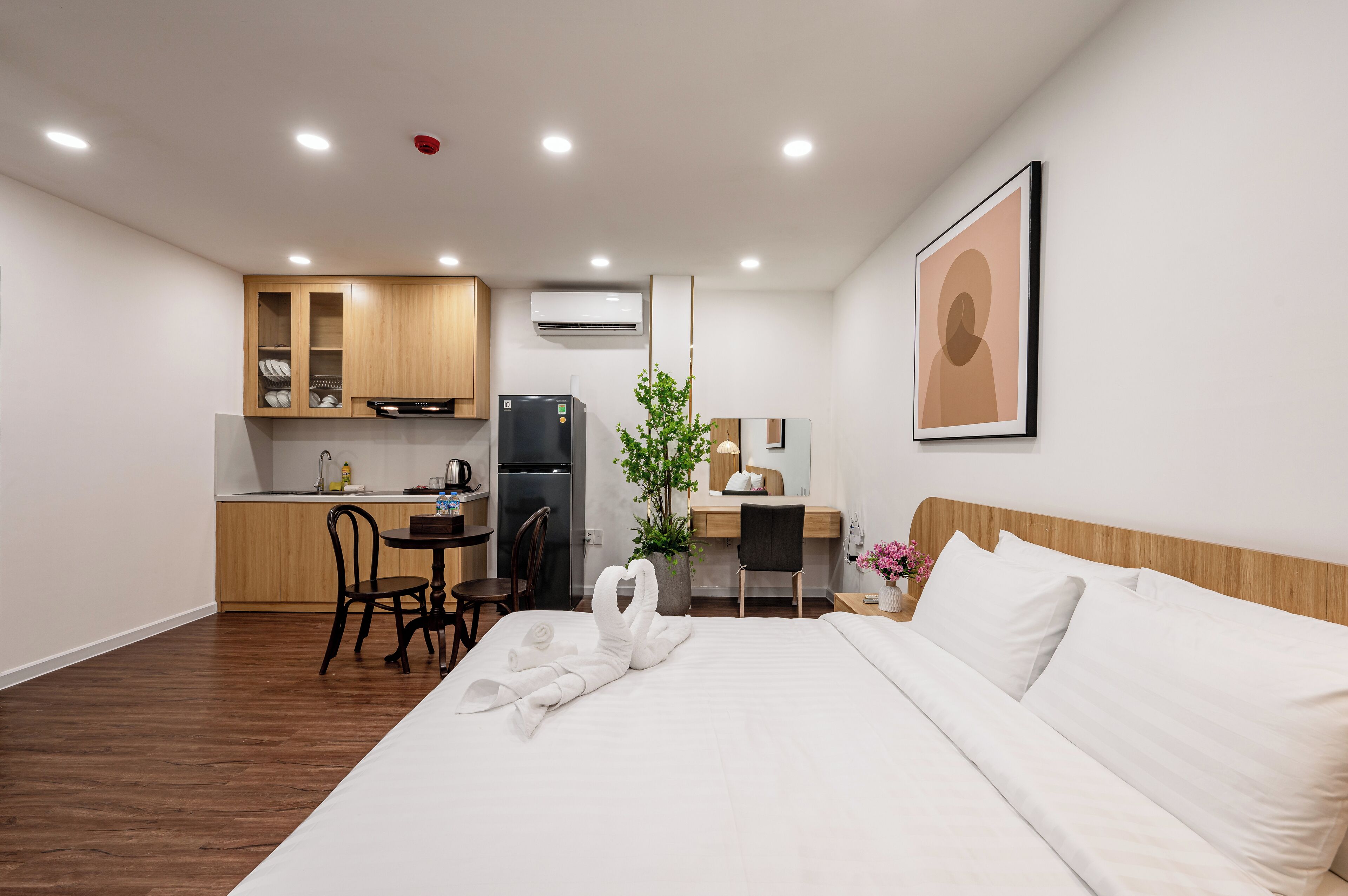 Habitació d'hotel amb llit, escriptori, cadira, zona de cuina petita i una planta en test.