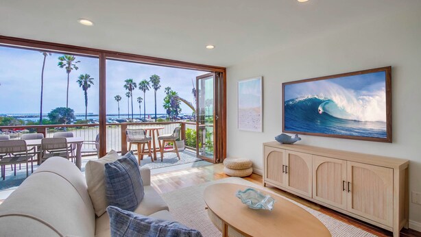 A spacious living area with a large window overlooking a balcony, a dining table, and a TV mounted on the wall. - Ocean Front * 3 Water View Patios * A/C (San Diego)