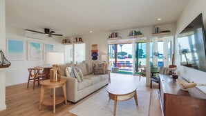 A spacious living room with a sofa, coffee table, and dining area. There are bookshelves and a large window providing a view of the outdoors. - Ocean Front * 3 Water View Patios * A/C (San Diego)