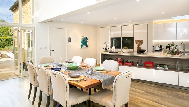 Cozinha moderna com armários brancos, sala de jantar com mesa e cadeiras de madeira, e vista para o exterior por uma porta de vidro.