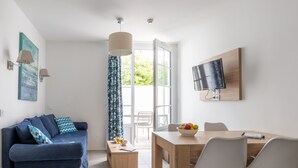 A modern living room with a blue sofa, a wooden dining table with chairs, a TV mounted on the wall, and a large window with curtains. - Résidence Odalys Collection Rève d'Ile by Interhome (Ile de Re)