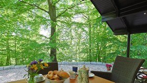 A dining table with a bowl of bread, a teapot, and a cup, set against a backdrop of a lush green forest.