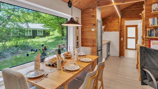 A wooden interior with a dining table set for four, a kitchen area, and a view of a green landscape.