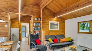 A cozy log cabin interior with a dining area, a sofa with colorful cushions, a glass coffee table, and a mounted picture.