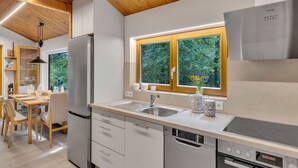 A modern kitchen with wooden ceiling beams, a built-in dishwasher, a stainless steel refrigerator, and a window with a view of greenery.