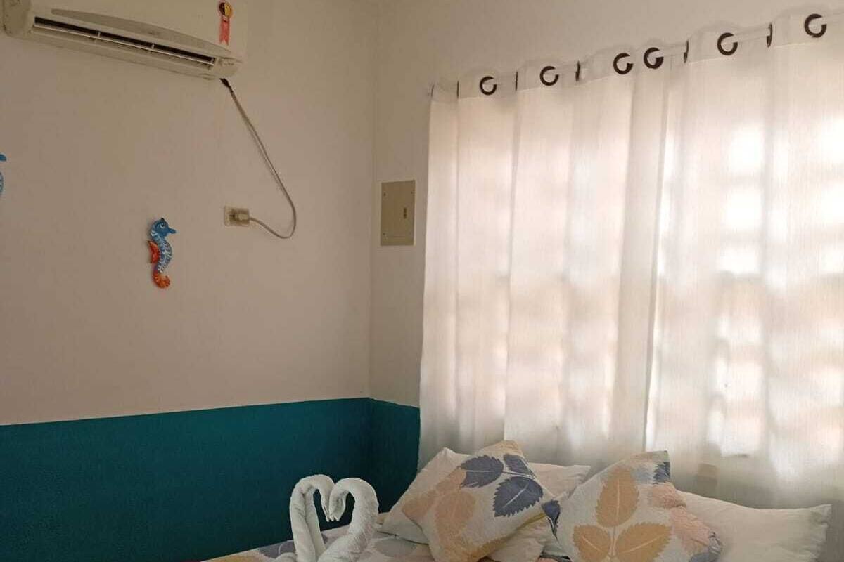 A bedroom with a bed, a red blanket, decorative pillows, a wall-mounted air conditioner, and a window with curtains.