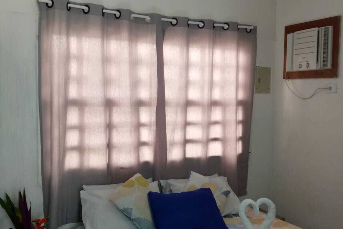 A bedroom with a bed, curtains, a wall-mounted thermostat, and a small plant.