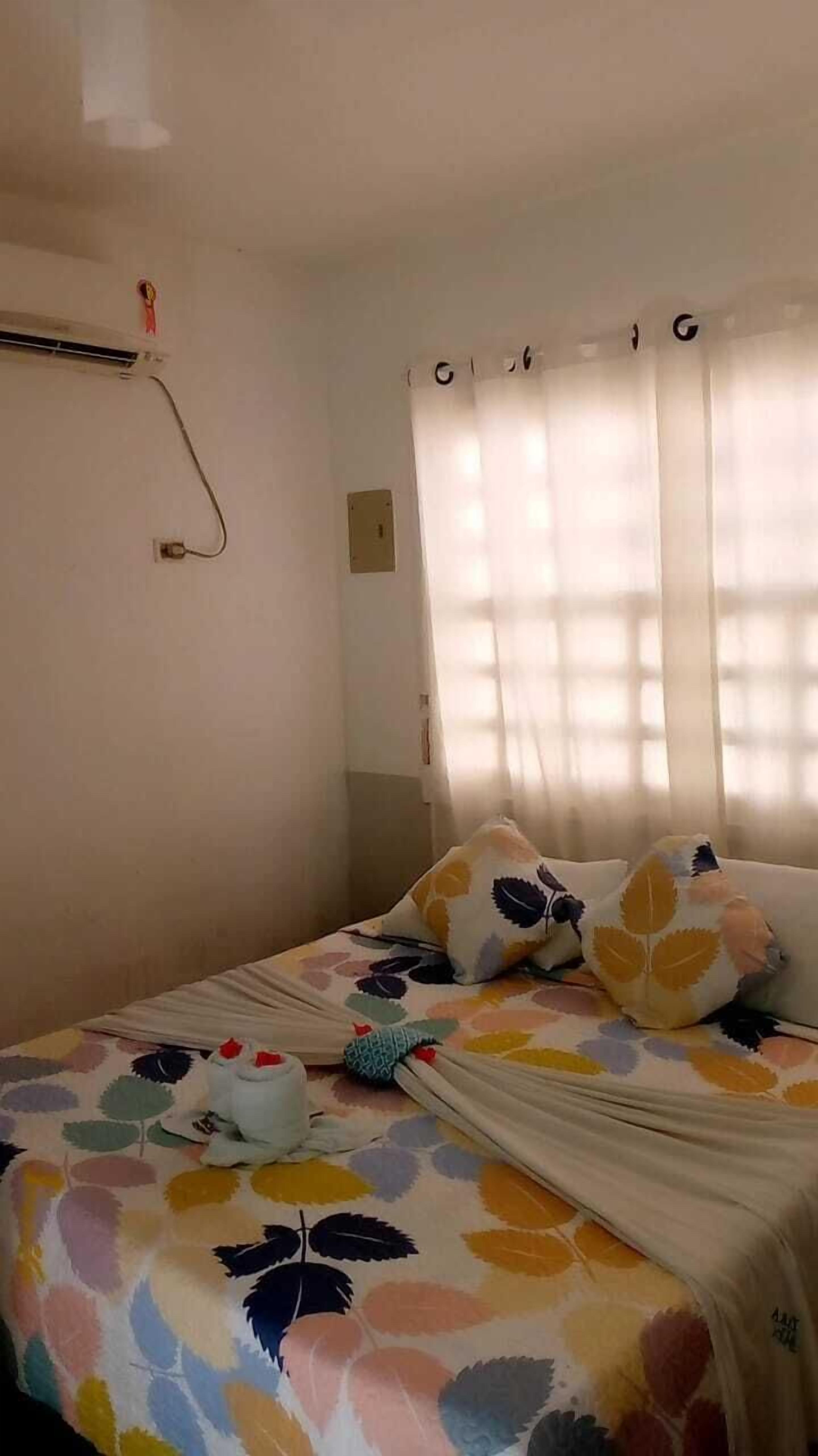 A bedroom with a bed, a window with curtains, a wall-mounted air conditioner, and a power outlet.