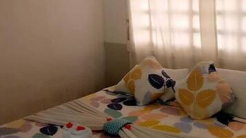 A bedroom with a bed, a window with curtains, a wall-mounted air conditioner, and a power outlet.
