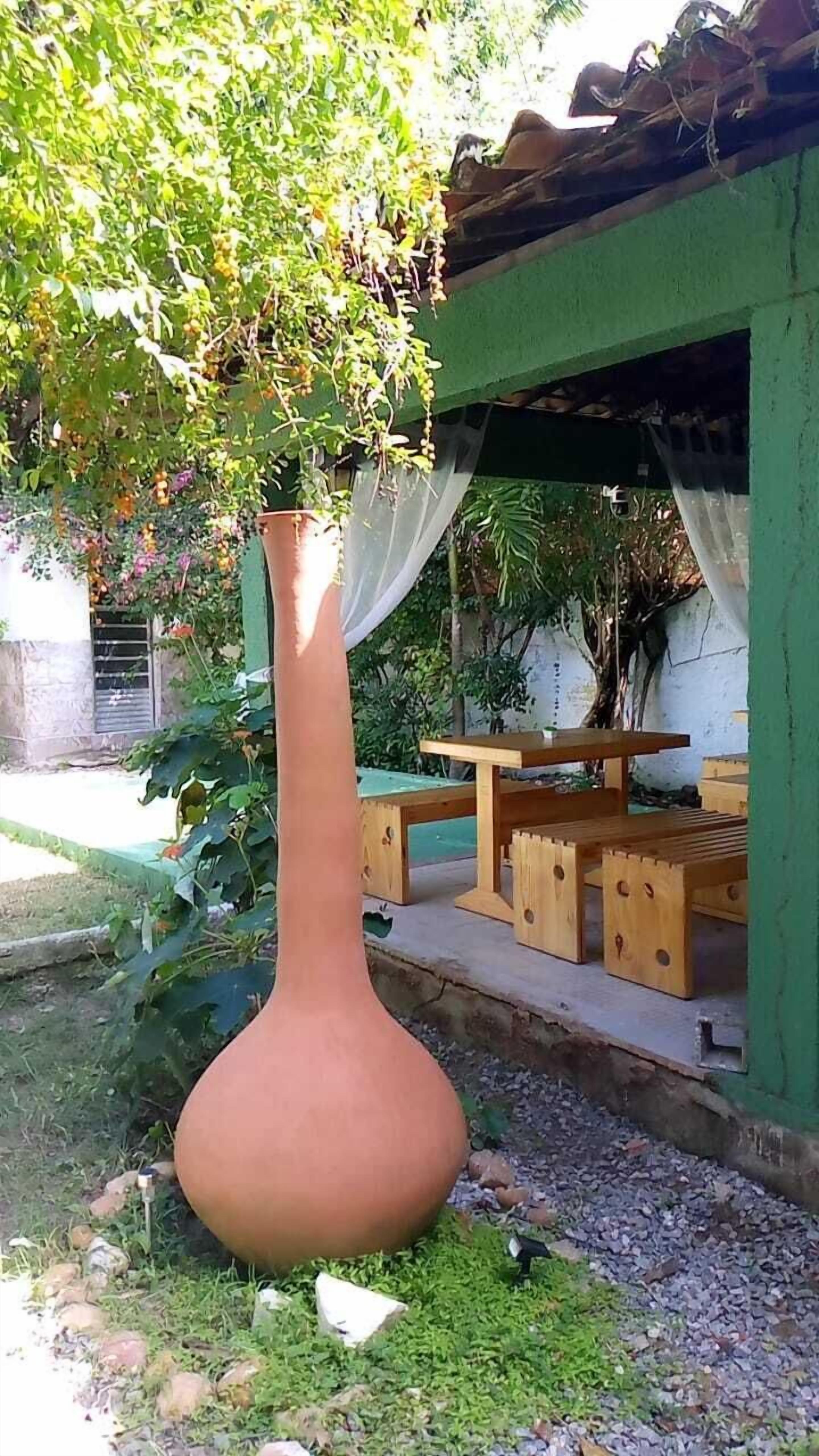 A large terracotta vase in a garden setting with a wooden table and chairs under a green structure.