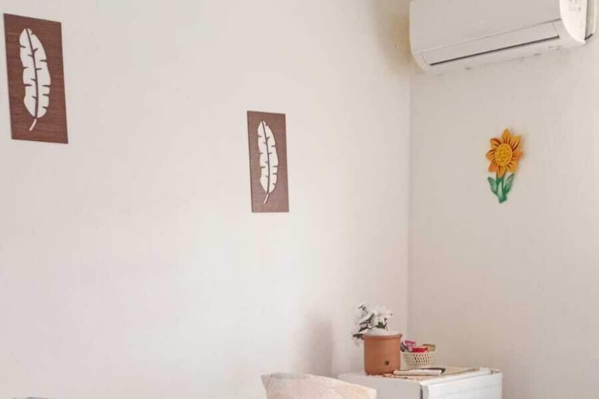 A neatly made bed with a leaf-patterned bedspread, pillows, and a folded towel. There is a small table with a plant and a basket, and an air conditioning unit on the wall.