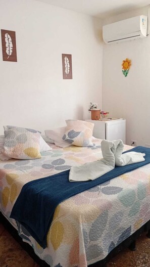 A neatly made bed with a leaf-patterned bedspread, pillows, and a folded towel. There is a small table with a plant and a basket, and an air conditioning unit on the wall. - Pousada La Floratta  (Paulista)