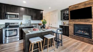 A modern kitchen with dark cabinets, stainless steel appliances, a large island with stools, and a built-in fireplace. - Downtown condo with a view and hot tub access (Bend)