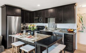 A modern kitchen with dark cabinetry, stainless steel appliances, a granite countertop, and bar stools. - Downtown condo with a view and hot tub access (Bend)