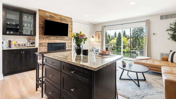A modern kitchen with dark cabinetry, a wooden accent wall, a flat-screen TV, and a dining area with a glass table and a sofa. - Downtown condo with a view and hot tub access (Bend)