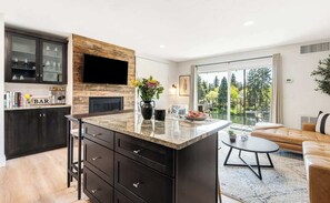 A modern kitchen with dark cabinetry, a wooden accent wall, a flat-screen TV, and a dining area with a glass table and a sofa. - Downtown condo with a view and hot tub access (Bend)