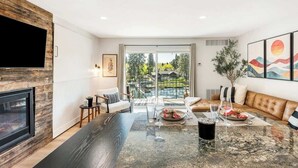 A modern living room with a fireplace, a dining area with a glass table, and a view of a green landscape through large sliding doors. - Downtown condo with a view and hot tub access (Bend)
