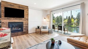 A modern living room with a fireplace, a flat-screen TV, a comfortable sofa, and a balcony with a view of trees. - Downtown condo with a view and hot tub access (Bend)