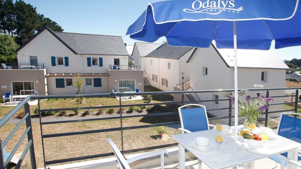 Ein Balkon mit Frühstückstisch, blauen Stühlen und einem blauen Sonnenschirm mit der Aufschrift 'Odalys'.