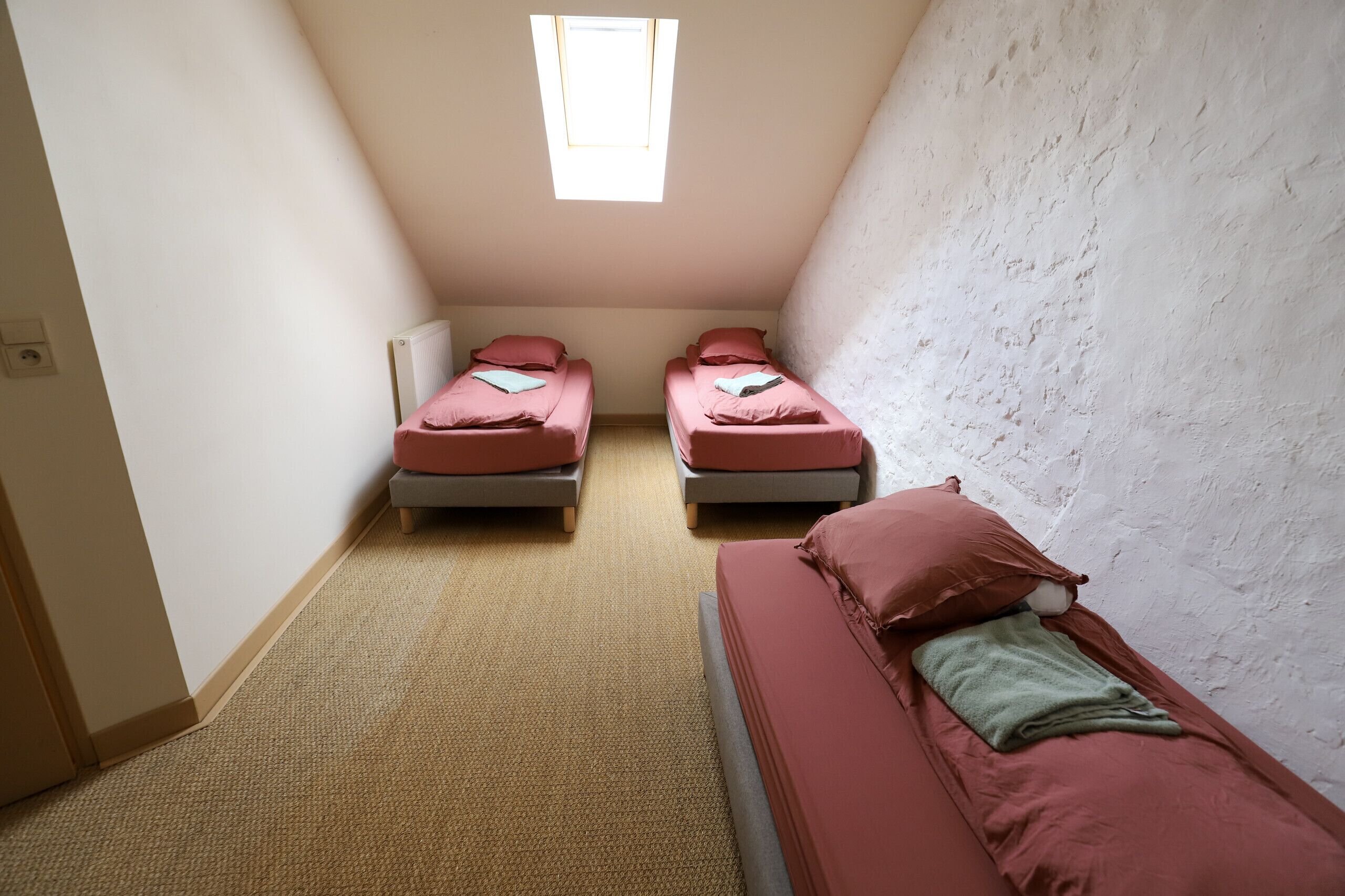 A small, narrow room with two beds and a third bed on the right. There is a window at the end of the room, and the walls are plain.