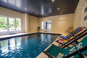 En inomhuspool med väggar av genomskinligt glas, ett tak med infälld belysning och en sittgrupp med solstolar.