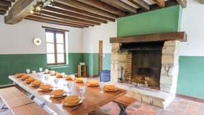 Una larga mesa de madera dispuesta con platos y copas de vino, una chimenea de piedra y una ventana con contraventanas de madera.