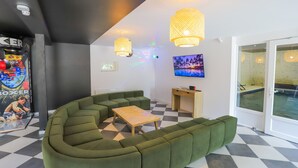 Un área de estar moderna con un sofá seccional verde, una mesa de centro de madera y un escritorio pequeño. Hay un ventanal con vista a una piscina, y la habitación tiene un piso a cuadros e iluminación colgante.