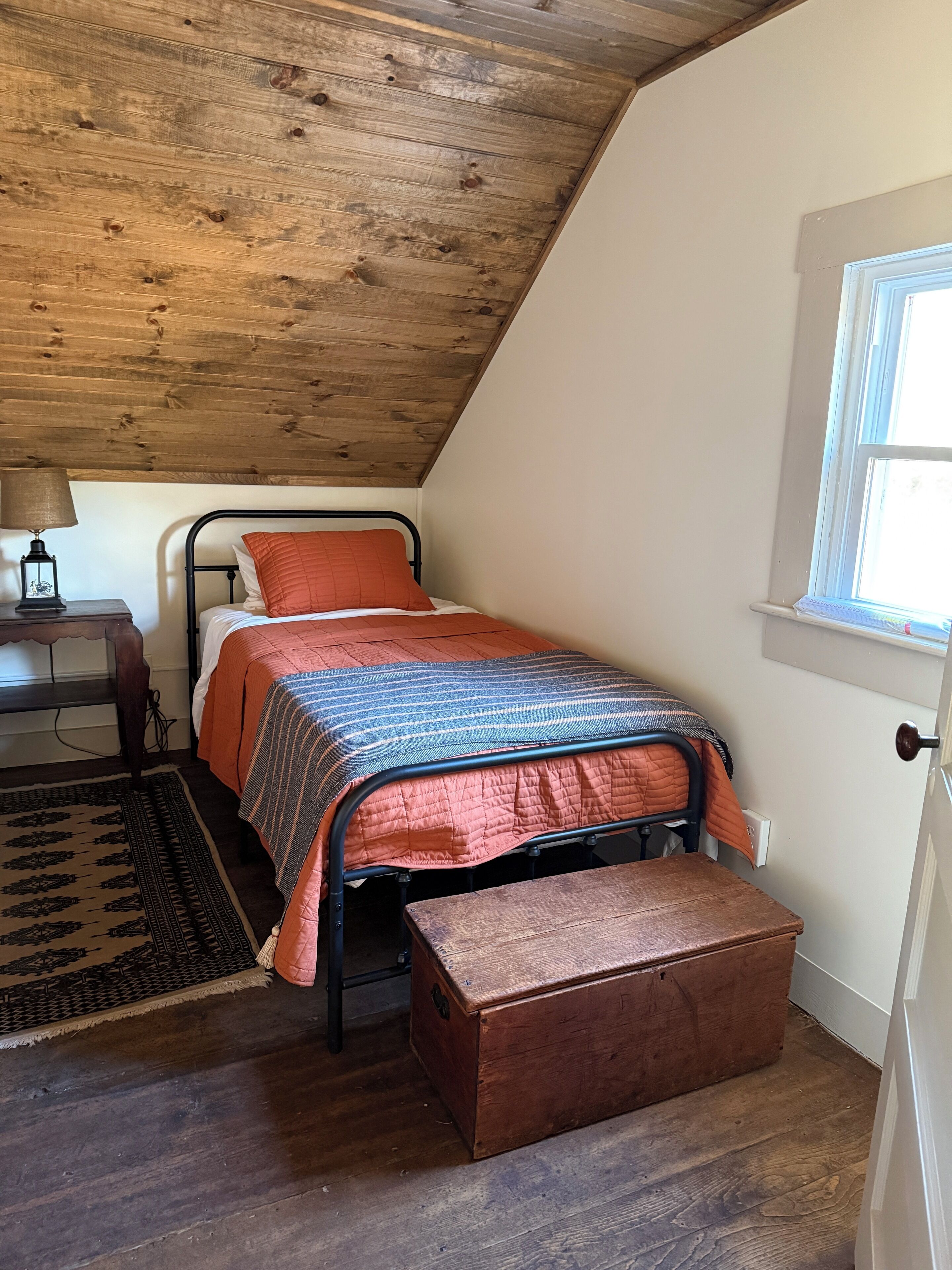 A bedroom with a sloped ceiling, a bed with a striped blanket, a wooden bench, a nightstand with a lamp, and a window with a view.