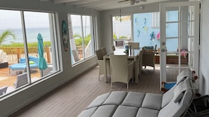A spacious room with a large window overlooking the ocean, a ceiling fan, and a dining area with wicker furniture. -  Cayman Bliss - Beachfront house private Pool & Beach Sleeps 8 Family-Friendly (West Bay)